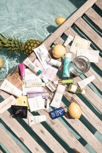 Flat lay of luxury skincare and sunscreen products on a wooden poolside deck with lemons, pineapples, and clear water in the background – includes brands like Coola, Ouai, Pixi, and Indie Lee, perfect for summer and spray tan-safe routines