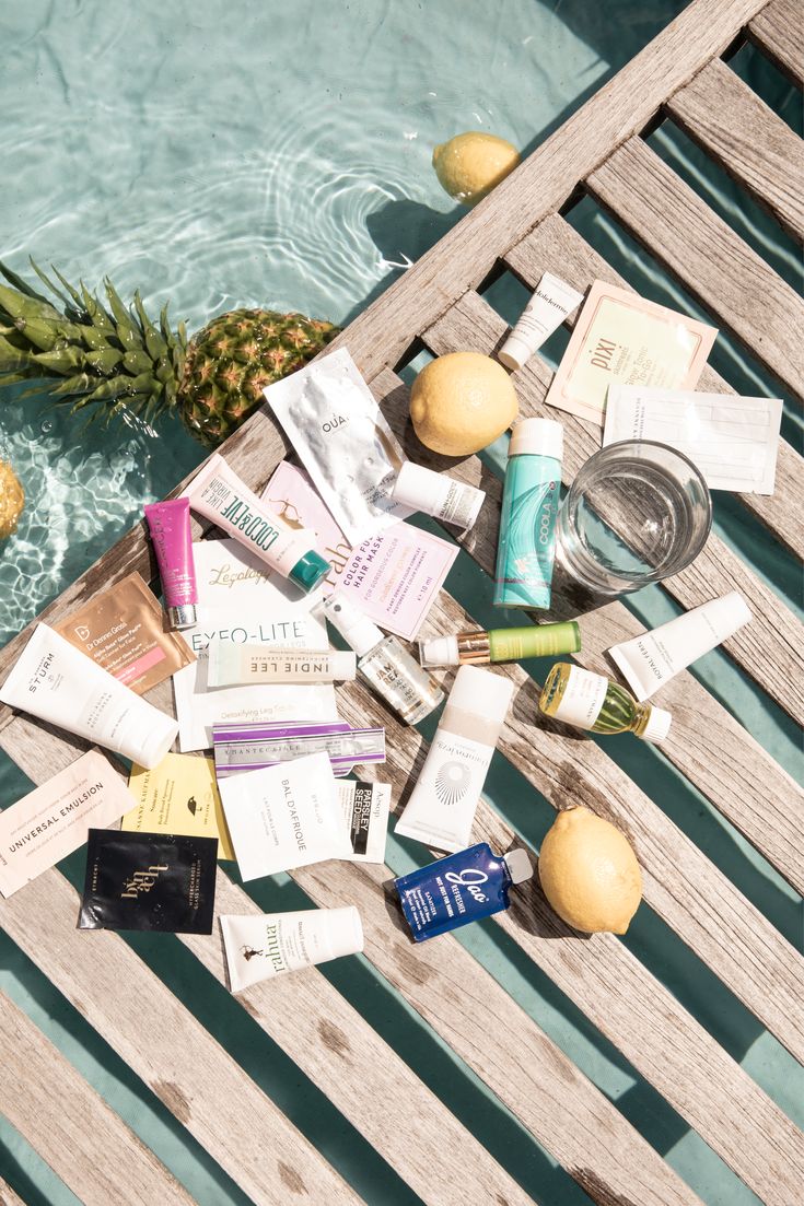 Flat lay of luxury skincare and sunscreen products on a wooden poolside deck with lemons, pineapples, and clear water in the background – includes brands like Coola, Ouai, Pixi, and Indie Lee, perfect for summer and spray tan-safe routines