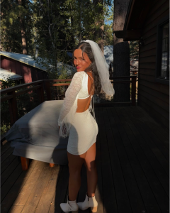 Bride in a white mini dress and veil with a natural spray tan glow on a wooden deck at sunset.